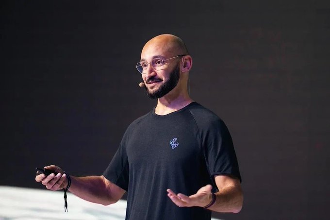 A bald, bearded man wearing glasses and a dark T-shirt speaks on stage with a headset mic and a presentation clicker in hand. The image appears to capture him mid-presentation about a stablecoin announcement, aligning with the post about discussing the new stablecoin alongside the team that powers it.