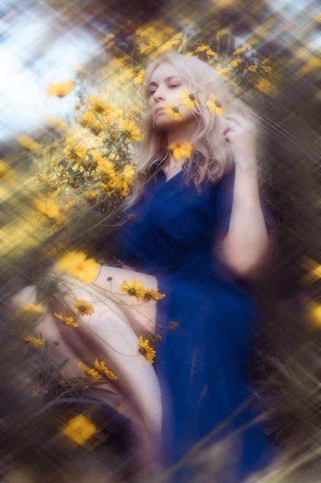 A woman with blonde hair in a blue dress sits in a field of yellow flowers with a dreamy, soft-focus effect.