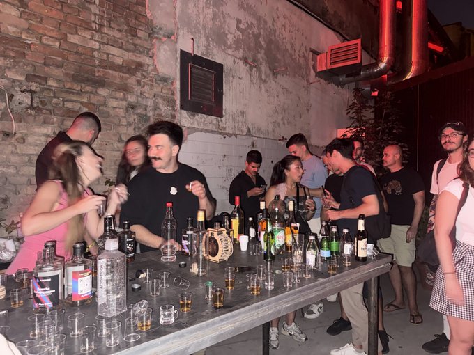 A lively bar or nightclub scene with a brick wall background, where a bartender in a black shirt serves drinks to a crowd of casual patrons gathered around a wooden counter covered with liquor bottles and shot glasses.