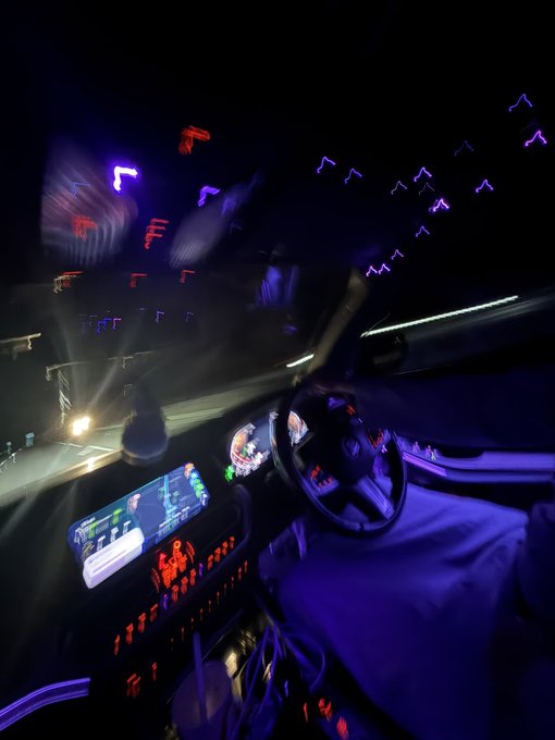 Interior of a vehicle at night featuring a vibrant, stylized LED ambient lighting setup with illuminated dashboard, steering wheel, and headliner displays showing animated icons, tied to the "GM bros" automotive customization community showcasing in-car lighting mods.