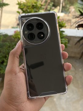A hand holds up a black OnePlus smartphone with a circular camera module in a clear case against an outdoor background with greenery.