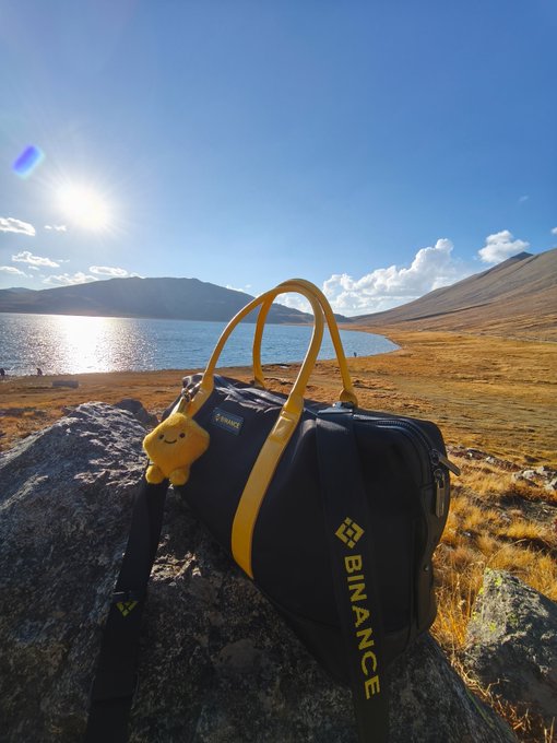 A navy and yellow Binance duffel bag, adorned with a small yellow plush charm, rests on a rocky outcrop overlooking a serene mountain lake under a bright sun, embodying the brand's global reach as its gear journeys to stunning, remote landscapes.