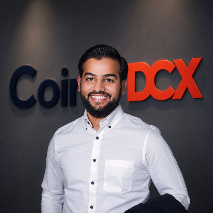 A smiling man in a white button-up shirt poses in front of a dark wall displaying the "CoinDCX" logo in black and red lettering, connecting to the post about CoinDCX's H1 2026 trading milestones of ₹14,600 crore in volume and 2.2 crore registered traders.
