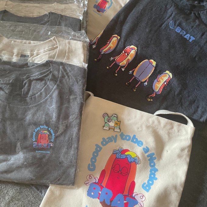 A collection of merchandise including t-shirts and tote bags featuring cartoon hotdog characters with the text 'good day to be a hot' and 'BRAT' printed on them.