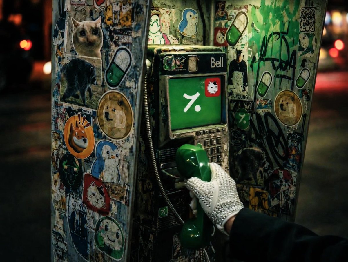 A hand wearing a white studded glove holds a green Bell telephone receiver at a graffiti-covered payphone with stickers and a green screen showing a percent sign.