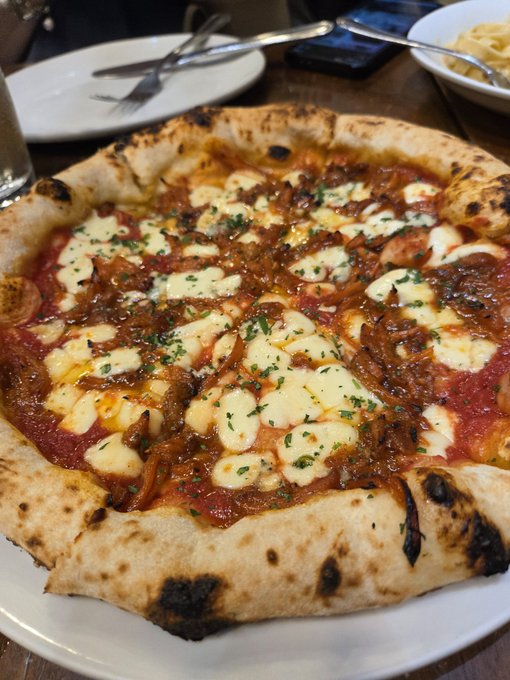 A freshly baked Neapolitan-style pizza topped with tomato sauce, melted mozzarella, caramelized onions, and parsley sits on a white plate, accompanied by utensils and a side bowl in the background, ready to be shared.