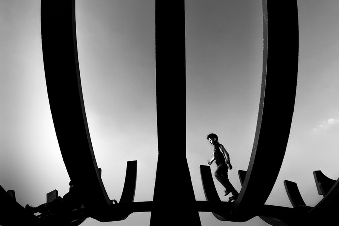 A silhouetted figure balances on a curved structure resembling cage bars or ribs, framed by the arching metal against a bright sky. The composition visually reinforces the artwork's title "Echoes of the Cage," suggesting entrapment, confinement, or the human condition within limiting structures.