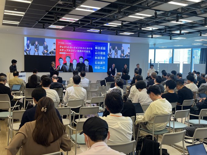 A large indoor conference or seminar in Japan, with an audience facing a panel of speakers seated at a table in front of a screen displaying Japanese text including 'スタートアップとビジネスにおける' and 'スタートアップコイン活用の可能性'.