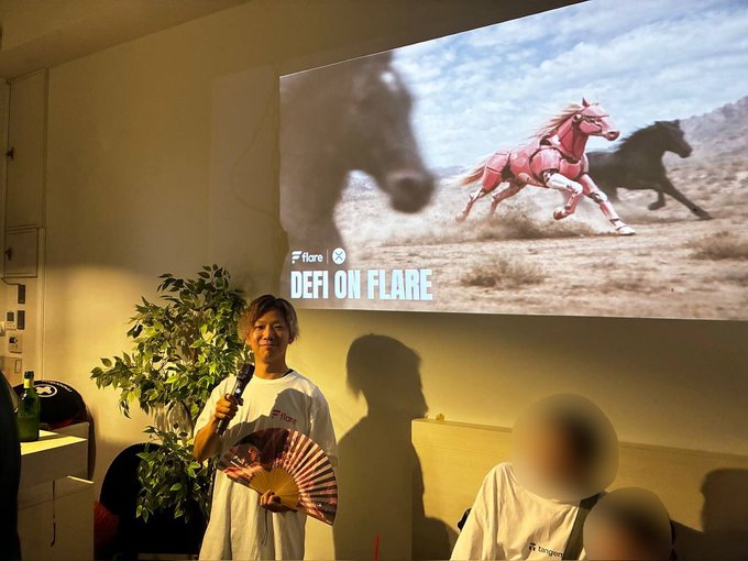 A woman stands holding a microphone and a decorative fan in front of a projected screen displaying "Flare DEFI ON FLARE" alongside an image of running horses, while attendees wearing Flare-branded shirts gather at what appears to be a Flare Networks crypto networking event.