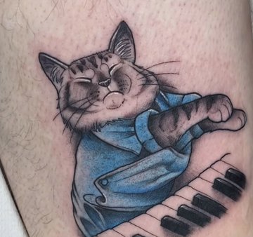 A tattoo on skin depicting a cartoon-style tabby cat with a content expression wearing a blue shirt, leaning against piano keys.