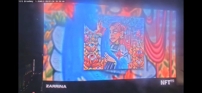 A billboard in Times Square displaying a colorful digital illustration of a woman surrounded by flowers and a butterfly, with the text "ZARRINA" and an "NFT NYC" logo.