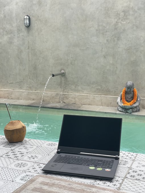 A laptop placed on a tiled patio near a pool with a Buddha statue and a flowing water spout in the background, with a coconut drink beside it.