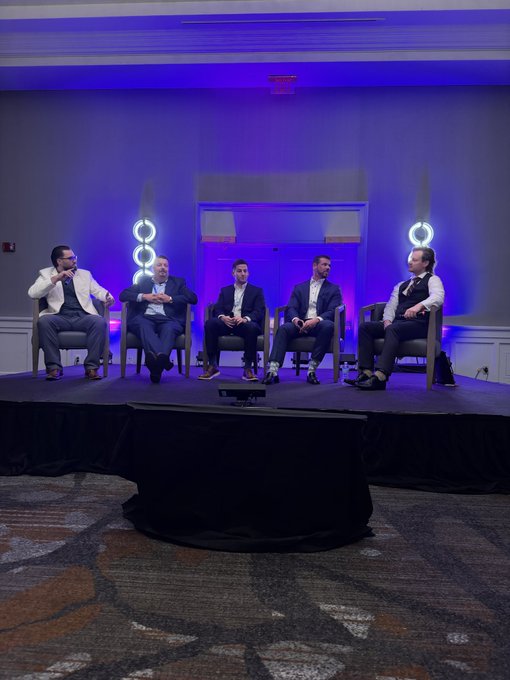 Five men in business attire sit on chairs arranged on a stage with blue and purple lighting and decorative ring-shaped fixtures, appearing to participate in a panel discussion. The scene reflects a professional conference setting, aligning with the Boston Blockchain Week event focused on insurance and wealth planning for digital assets.