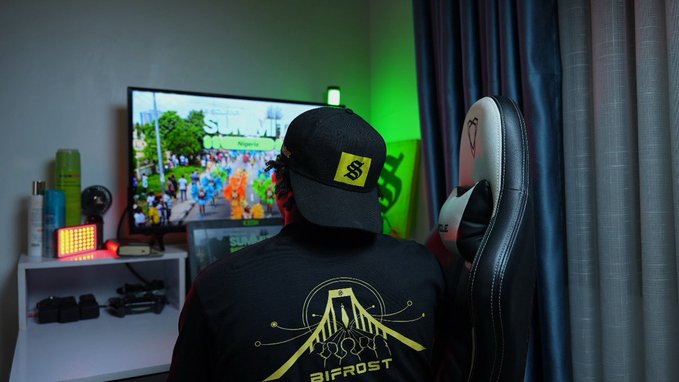 A person wearing a black "Bifrost" shirt with a yellow logo and a black cap with a yellow "SB" patch sits in a gaming chair facing a monitor displaying "SolAi." The image showcases the wearable design created for the @SolflareBifrost team, reflecting the brand's aesthetic.