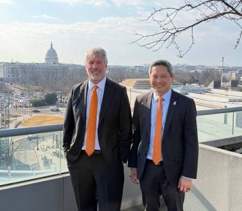 The image shows two men wearing suits, one with an orange tie and the other with a white shirt and black suit. They are standing next to each other on a balcony or deck overlooking a cityscape. The man on the left has his hand in his pocket while the man on the right is holding onto his tie. Both of them have their hands visible in the image, which might suggest that they are posing for a photo together.