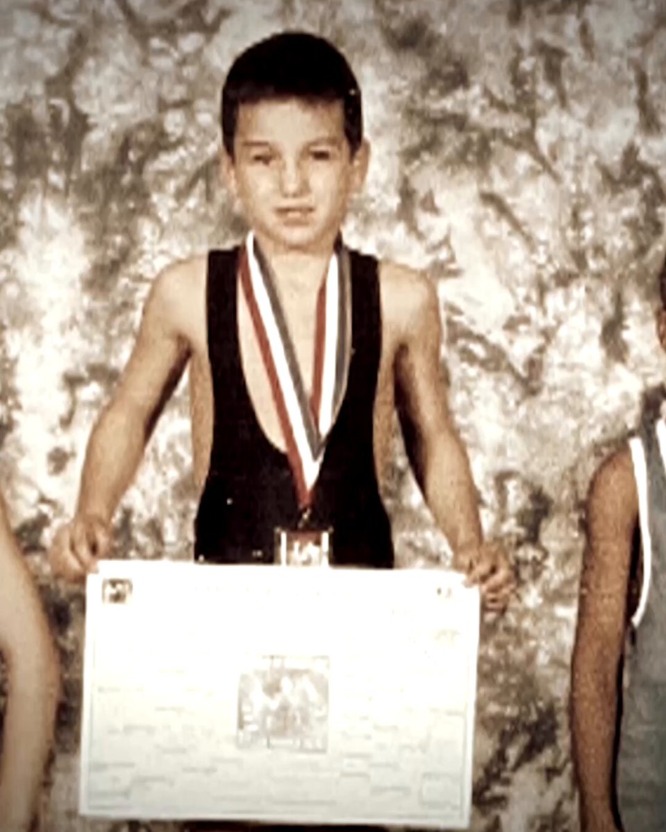 The image features a young boy holding up a certificate or award, possibly for his wrestling achievements. The background of the photo has a greenish hue with white splotches, giving it an old-fashioned appearance. There are two other people present in the scene, one on each side of the boy, but they don't seem to be directly involved with the main subject.