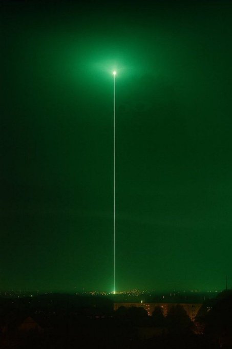 A bright green laser beam shoots straight up into the night sky from a distant cityscape, creating a glowing green halo.