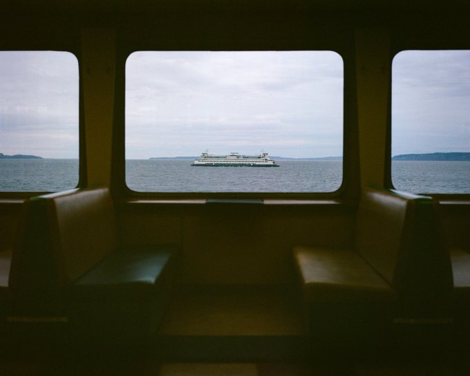 A view from inside a ferry cabin, with empty cushioned seats in the foreground framing a window that looks out onto another passenger ferry crossing a calm body of water under a cloudy sky. The image complements the "Available 1/1 works on @SuperRare" post by serving as the NFT artwork being offered for sale on the platform.