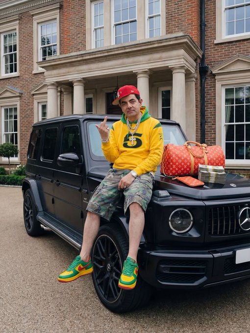 A man wearing a yellow hoodie, camouflage shorts, a red cap, and green and yellow sneakers sits on the hood of a black Mercedes-Benz G-Wagon parked in front of a large brick mansion, with a red Louis Vuitton duffel bag on the vehicle beside him.
