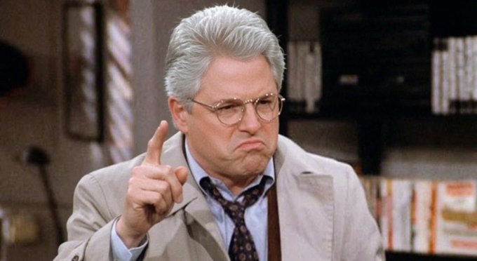 A man with gray hair and glasses, wearing a beige jacket and patterned tie, pointing his finger upward with a disapproving expression in what appears to be an office setting.