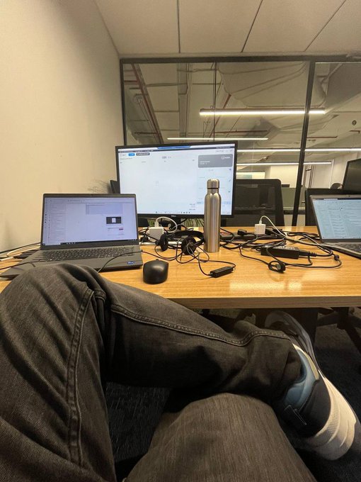 A casual first-person view shows someone's legs in dark jeans and sneakers stretched out under a wooden desk cluttered with a laptop, an external monitor, water bottle, tangled cables, and small accessories in what appears to be an empty office space.

The image connects to the post by illustrating the quiet, solitary work environment of someone independently building a project ("HACD Labs"), with the empty office underscoring the focus on long-term development while an established community member shows unexpected early support.