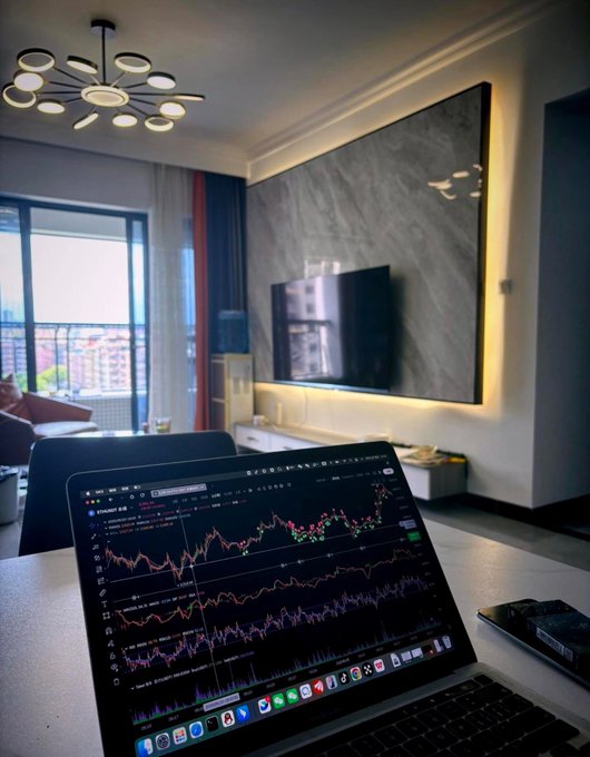 A laptop displaying colorful trading charts sits on a desk in a modern apartment, with a marble-patterned TV wall, a chandelier, and a balcony view of city buildings in the background.