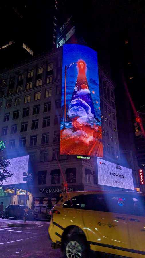 A nighttime city street scene featuring a large curved digital billboard displaying a colorful cosmic/space scene above Cafe Manhattan, with a yellow taxi in the foreground and a Sparky store visible.