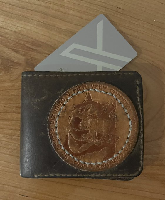 A brown leather bifold wallet containing a custom copper-colored coin or medallion engraved with a cat face and German text "Crasio DIE Katze w rielt vera n aranch xamal bin not mo Sostosa", with a white card sticking out.