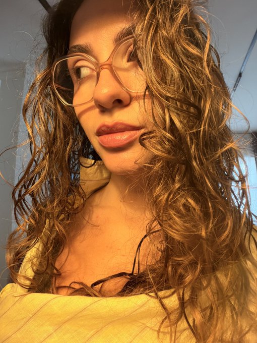 Close-up selfie of a young woman with long curly brown hair wearing orange-tinted eyeglasses and a yellow top, lit by warm sunlight.