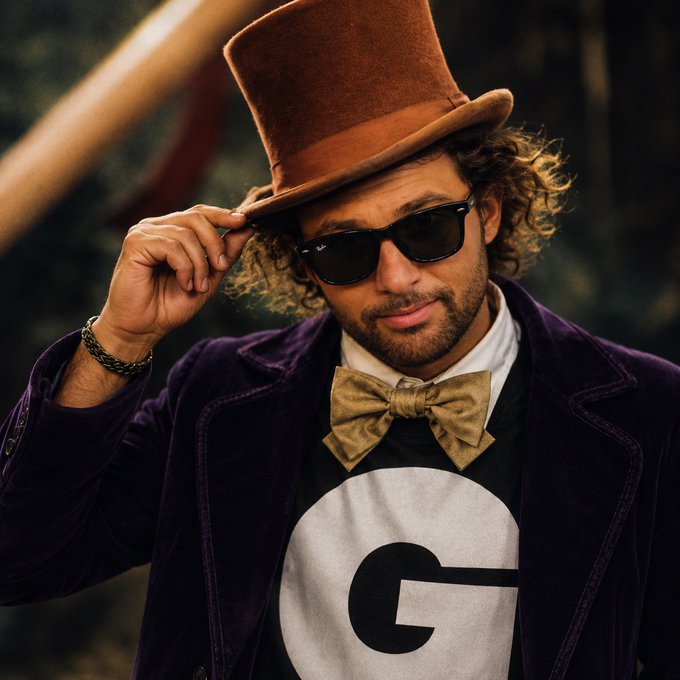 A man with curly hair wearing a brown top hat, sunglasses, a purple velvet jacket, a beige bow tie, and a shirt with a large 'G' logo, reminiscent of Willy Wonka.