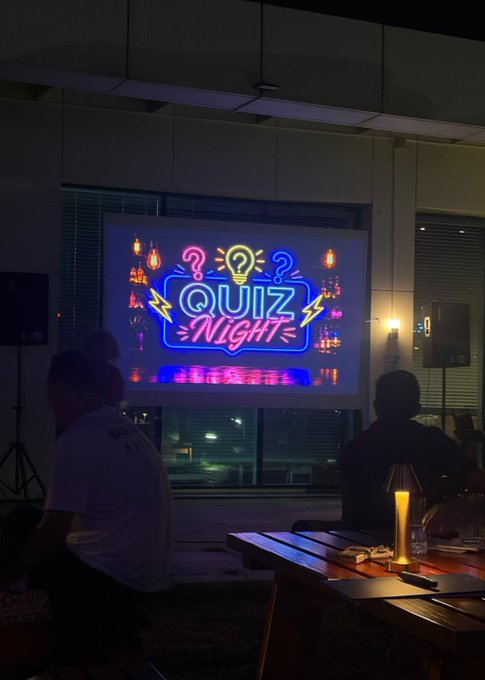 A neon 'QUIZ NIGHT' sign projected on a screen in a dimly lit venue with people seated at tables.