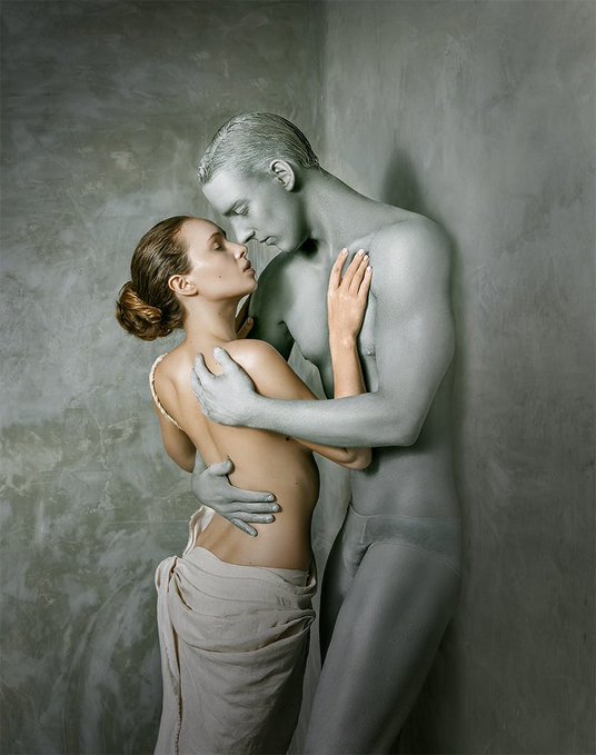 The image features a man and woman embracing each other, with the woman's body partially covered by her shirt or skirt. The couple appears to be in love, as they are standing close together against a gray wall backdrop. The photo captures their intimate moment, conveying a sense of closeness and affection between them.