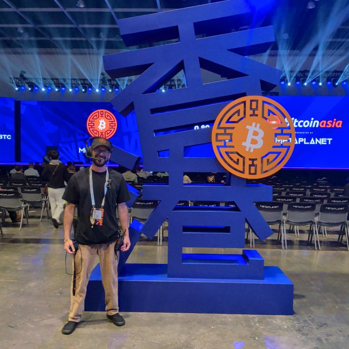 A man wearing headphones and a cap stands in front of a large Bitcoin symbol installation at what appears to be a Bitcoin Asia conference event, with seating and stage lighting visible behind him.