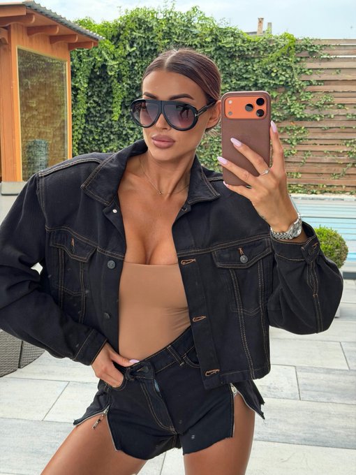 A mirror selfie of a woman wearing dark sunglasses, an open black denim jacket over a beige top, and denim shorts, posing outdoors near a pool area.