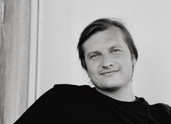 A black and white portrait photograph of a smiling young man with shoulder-length hair wearing a dark shirt against a plain wall background.