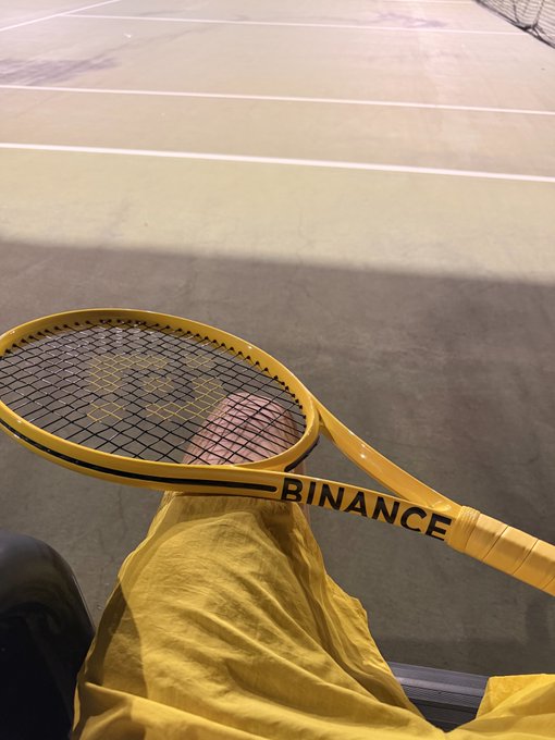 A yellow tennis racket with 'BINANCE' written across the strings and shaft, resting on a person's yellow-clad lap on a tennis court at night.