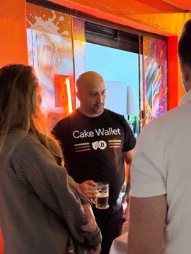 A man wearing a black 'Cake Wallet' t-shirt holding a drink stands between a woman with blonde hair and another man, seemingly at an indoor social gathering.