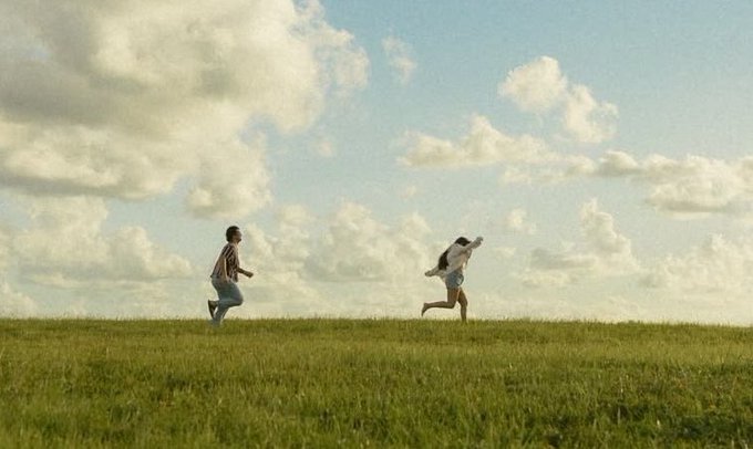 Two children running joyfully across a grassy field under a partly cloudy blue sky in a cinematic, nostalgic photograph.