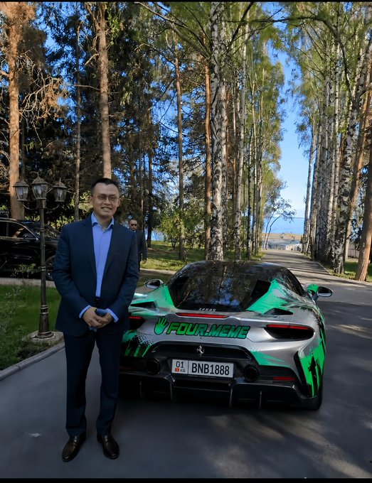 A man in a dark suit and glasses stands next to a green and silver Ferrari sports car with 'FOUR.MEME' branding. The car bears the license plate 'BNB1888', and the setting appears to be a tree-lined driveway.