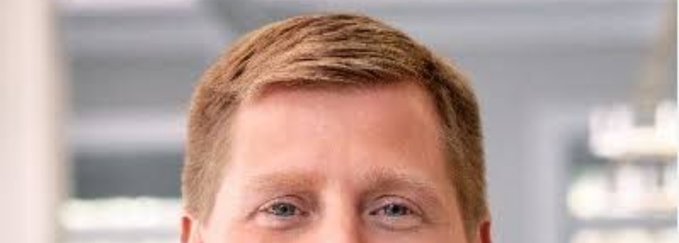 A close-up headshot of a fair-haired man smiling, with a blurred indoor background.