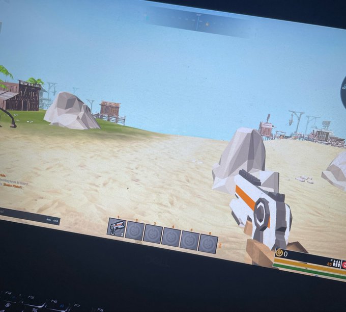 A low-poly first-person shooter game is displayed on a Dell laptop screen, showing a sandy beach environment with rocks, distant structures, and a weapon inventory bar at the bottom.