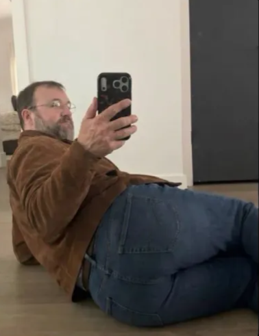 A bearded man wearing glasses, a brown jacket and blue jeans lies on a floor while holding up a smartphone to take a photo or selfie.