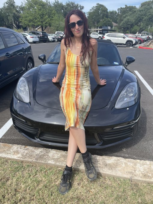 A woman in a striped orange and yellow dress leans against a black Porsche 918 Spyder in a parking lot, fitting the post's claim that "Dogecoin gets you a hot girlfriend and a Porsche confirmed." The luxury sports car and stylish companion serve as visual evidence—though humorous—of cryptocurrency success.