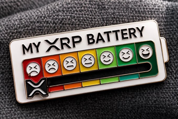 A close-up of a rectangular enamel pin on a textured gray fabric, displaying a battery-style gauge with emoji faces and the text 'MY XRP BATTERY' along with an XRP logo.