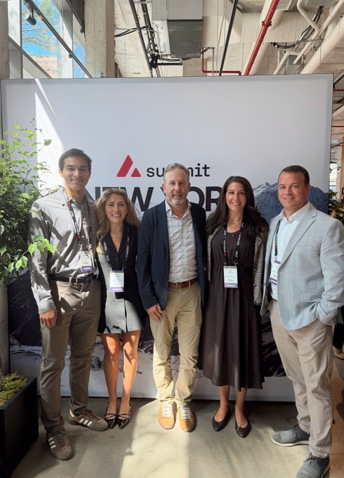 Five people pose together in front of a Summit New York event banner, with attendees wearing lanyards and business casual attire.