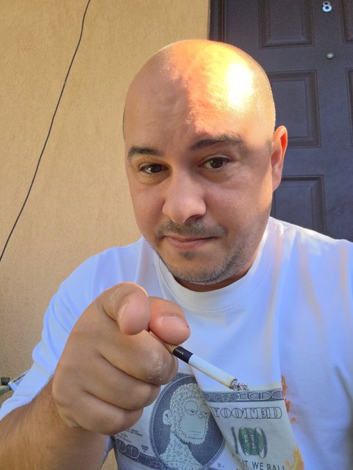 A bald man with a goatee in a white t-shirt featuring a hundred dollar bill graphic points a cigarette toward the camera.