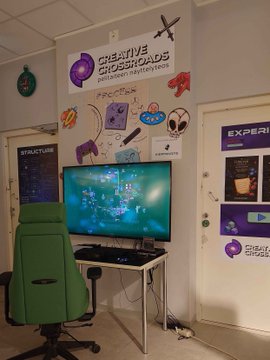 A gaming setup featuring a green racing chair, a monitor displaying a game, and colorful wall decorations including a "CREATIVE CIRCUS ARCADE" banner with various gaming-themed stickers like rockets, controllers, and UFOs.