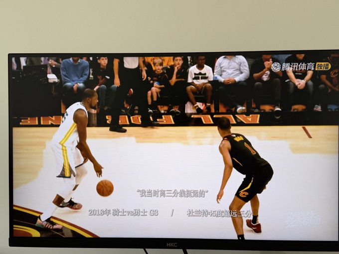 The image shows a basketball game being played between two teams, with one team wearing yellow jerseys and the other wearing black jerseys. The players are actively engaged in the game, with one player holding a basketball while another player is dribbling the ball on the court. A large TV screen displays the ongoing match, capturing the attention of the audience who can be seen watching from various positions around the room.