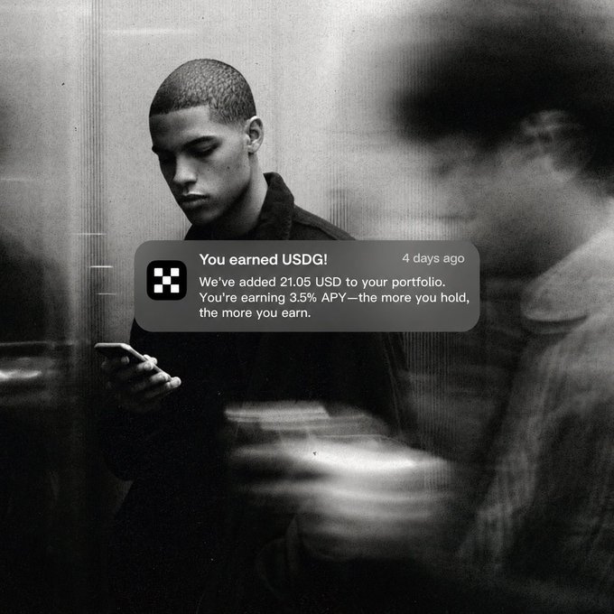 A young man looks down at his phone while another person rushes past him in a blurred black-and-white scene, as an overlay notification reads "You earned USDG! We've added 21.05 USD to your portfolio. You're earning 3.5% APY—the more you hold, the more you earn." The image reinforces the post's message by visually capturing a moment of everyday life where passive income quietly accrues in the background.