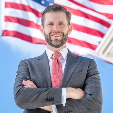 Eric Trump stands smiling with arms crossed in a gray suit and pink tie, posed in front of an American flag. The patriotic backdrop visually reinforces the "American Bitcoin" branding tied to his company's upcoming Q2 2026 earnings release on August 3.